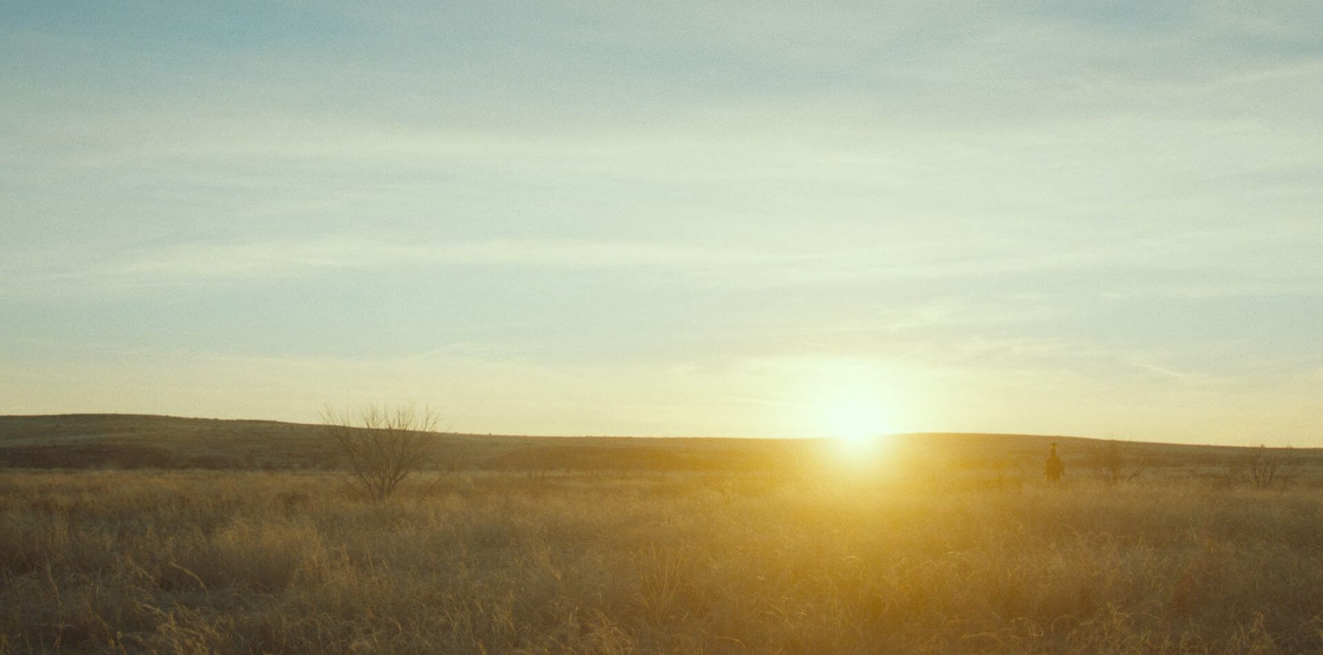 Open prairie at sunrise, the sun low on the horizon and a figure far off in the distance.